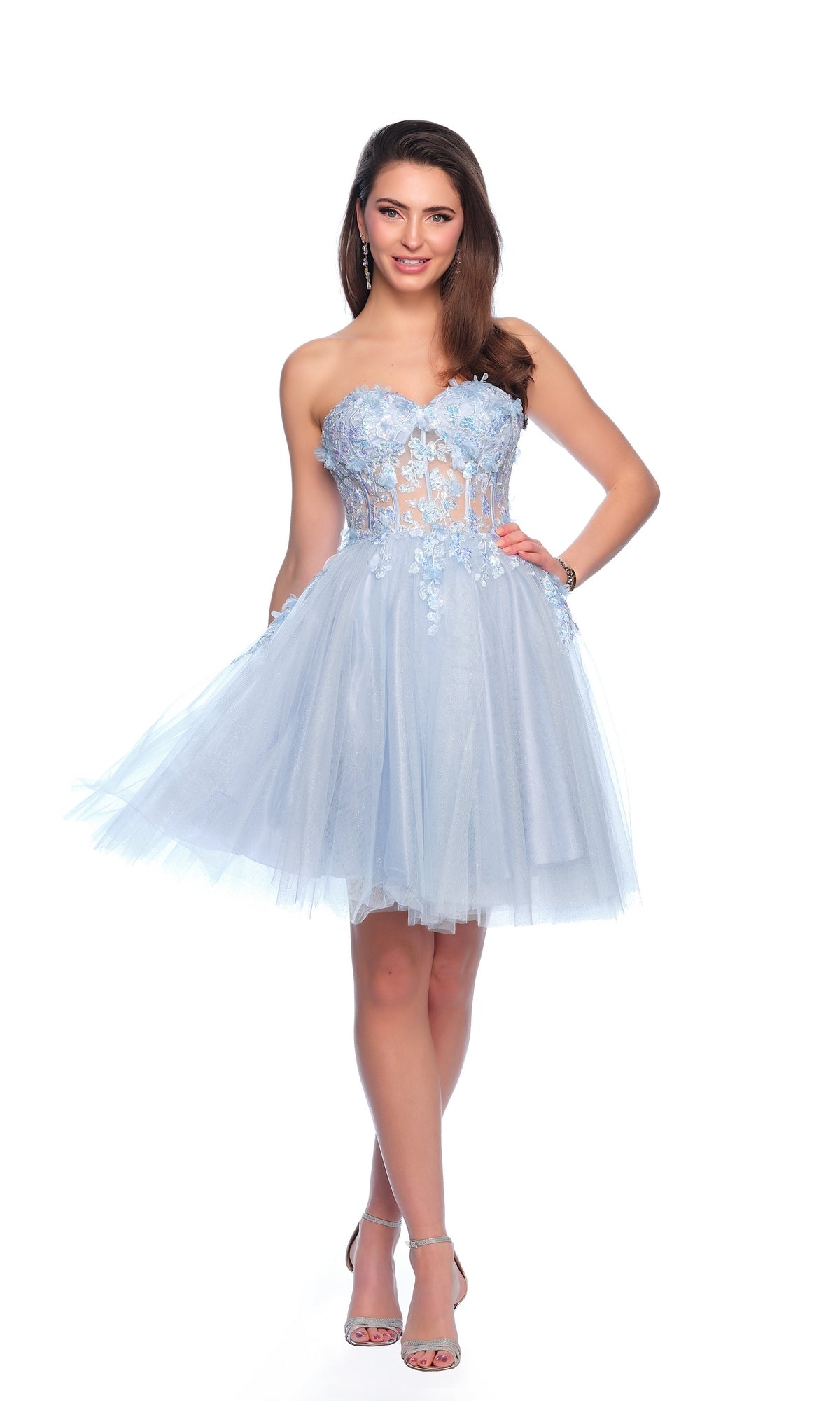 Woman wearing the Sheer Glitter Homecoming Dress 11973—a light blue, strapless, knee-length dress with floral embroidery and a sheer bodice—stands smiling against a white background.